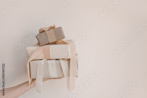 Birthday, Christmas gift concept. Female hand holding stack of gift boxes with beige ribbons over white wall. Holiday background with blank copy space