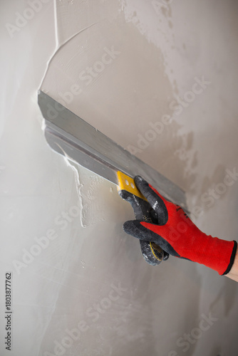 A masters hand in a red glove with a spatula does the work of leveling a wall with putty.