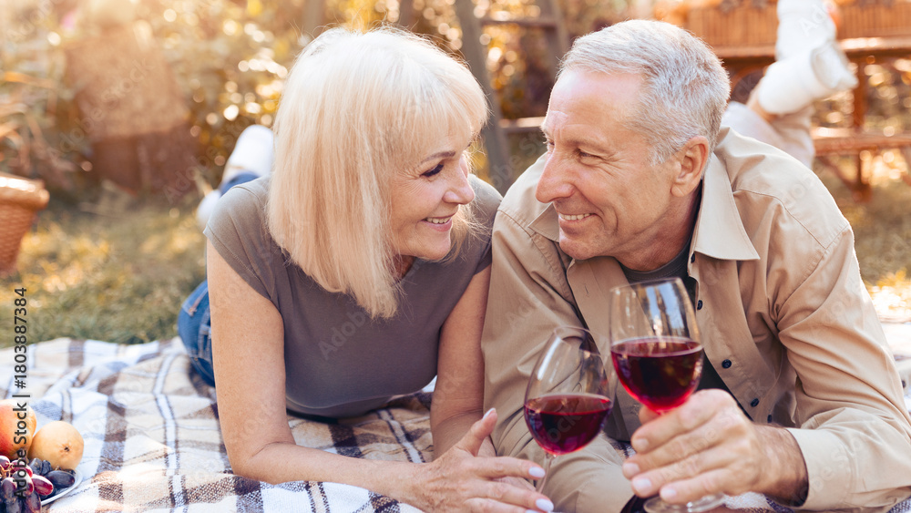 Fototapeta premium A happy couple lies on a blanket in a garden, sharing a joyful moment over glasses of red wine. They smile at each other, surrounded by fruits and a peaceful outdoor setting.