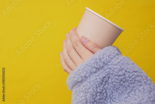 Craft eco friendly disposable cardboad cup in a female hands wearing cozy sherpa sweatshirt. Yellow background. Coffee or tea drink