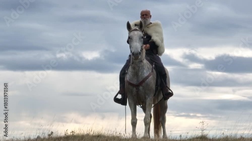 Aerial drone establishing or tracking shot of Bearded warrior in fur cloak riding a white horse across open landscape at sunrise, mountains in the background. Historical, rugged, adventurous scene