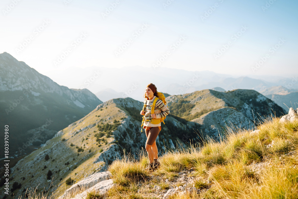 Fototapeta premium Beautiful female hiker with yellow backpack stands on mountain ridge, watching sunset. Young woman enjoys journey and feels freedom of sunset. Concept of hiking, freedom, and an active lifestyle.