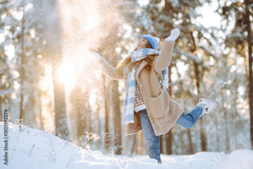 A smiling woman is having fun in a snowy forest. A sweet woman enjoys a winter day, throwing snow in a sunny park. Concept of a walk, cold.