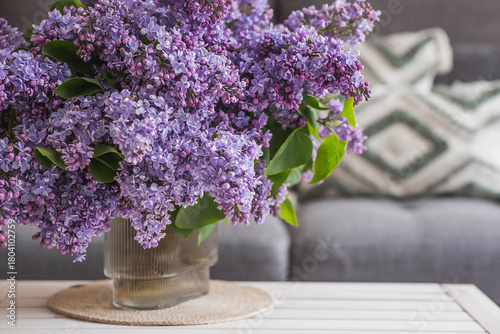 The concept of spring, home comfort, and celebration. A large purple bouquet of lilacs on the table. In the background, the interior of a modern living room.
