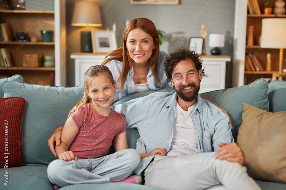 Obraz premium A smiling couple and their daughter sit together on a soft couch, sharing joyful moments in a warm and inviting living room filled with natural light.
