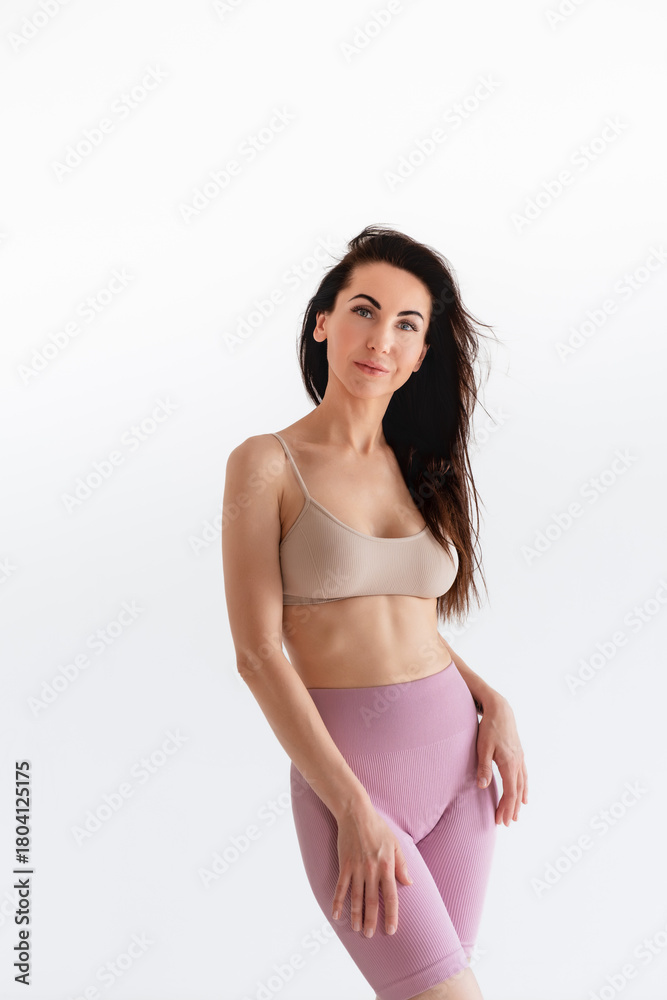 Fototapeta premium Sports woman standing in activewear with abs muscles, looking at camera isolated on white background. Caucasian 30s fitness girl wears beige top, pink leggings. Sexy fit smiling brunette, blue eyes.