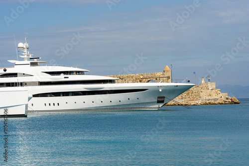 Luxury white superyacht moored by historic stone fortress in calm turquoise sea, symbol of wealth, tourism and nautical lifestyle under soft cloudy sky.