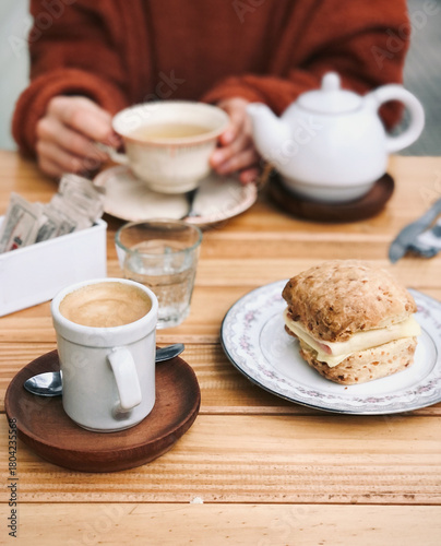 Cozy close-up shot of an outdoor breakfast or brunch setting with a coffee cup, a water glass, a savory scone sandwich, and a person holding a teacup and teapot on a wooden table.