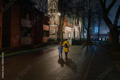 Mother and daughter walking at night toward an illuminated brick church in Kaunas, a festive winter street with wet reflections.