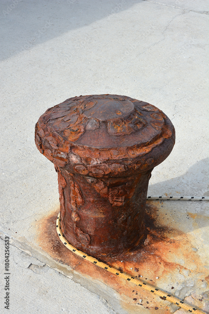 Fototapeta premium A heavily rusted, cylindrical metal mooring bollard stands on a concrete pier, with a thick, frayed rope looped around its base