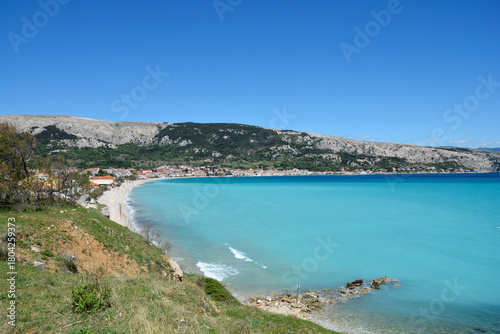 Fototapeta Naklejka Na Ścianę i Meble -  A panoramic view captures the crescent-shaped Vela Plaza beach and the town of Baska on Krk Island, with turquoise sea and a dramatic mountain backdrop under a clear sky