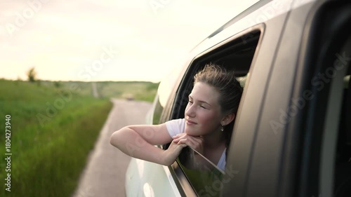 Young girl leans from car window looking toward winding road through green grass and countryside portrait of teenager during travel summer light outlines braid and thoughtful expression car window
