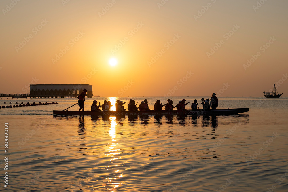 Naklejka premium Doha, Qatar - October 24, 2025: Silhouette people rowing dragon boat at sunset time Katara Cultural Village 