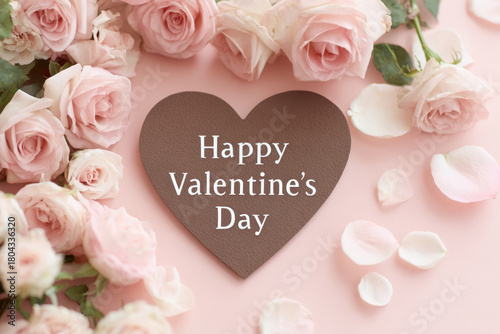 Chocolate paper heart card surrounded by pale pink roses and scattered rose petals on soft blush background conveying romantic celebration and gentle affection