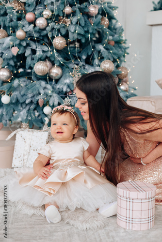 Family celebration with a little girl in a fancy dress beside a decorated Christmas tree and a joyful woman during a festive gathering
