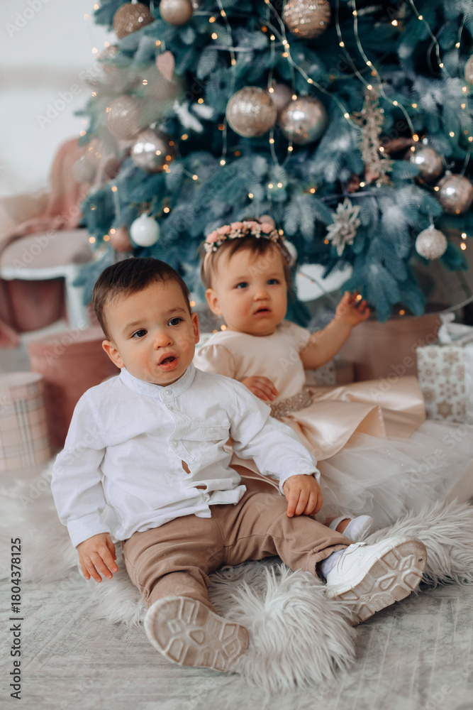 Fototapeta premium Children sitting on a soft rug next to a decorated Christmas tree in a cozy indoor space filled with holiday decorations and gifts