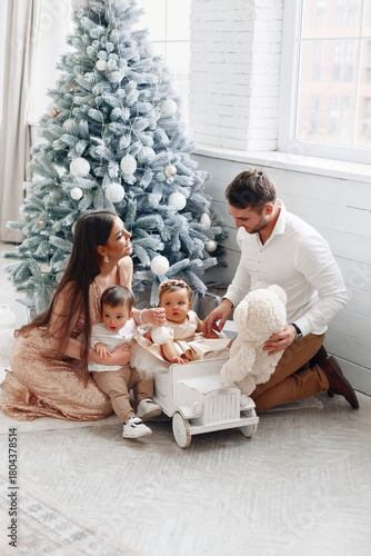 Family of four enjoys a cozy holiday moment by the Christmas tree with their toddlers playing with a vintage toy