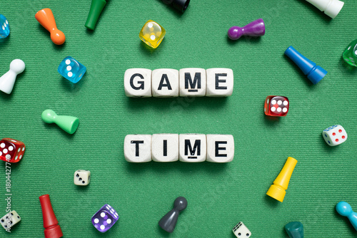 Wooden dice show 'Game Time' with a selection of colourful dice and game tokens lying around it on a vibrant green baize fabric