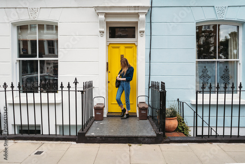 A young woman stands on a doorstep in front of a vibrant yellow door, holding her bag. The colorful building is located in the London Borough of Camden.