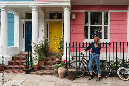 A young woman in jeans and a blue shirt stands with her bicycle in front of colorful houses with potted plants on the steps in London Borough of Camden.