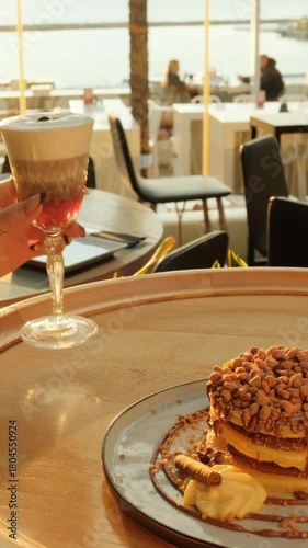 A hand holds a layered dessert coffee in warm sunset light beside a plate of stacked pancakes with nuts, captured in a cozy outdoor café setting.
