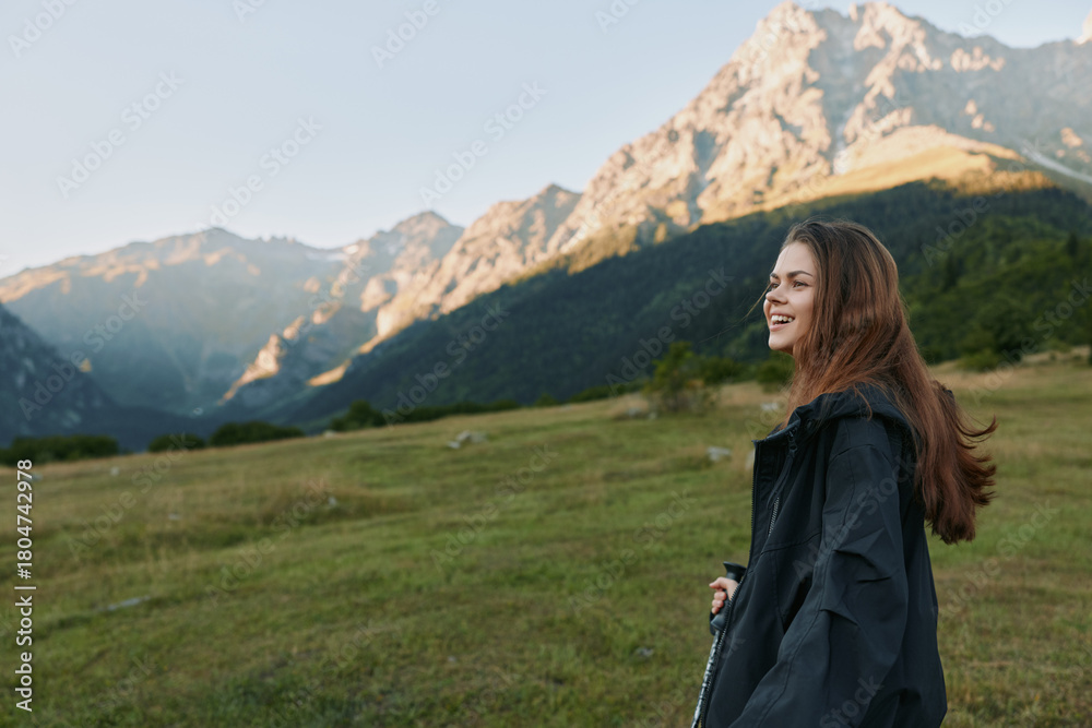 Fototapeta premium A smiling woman stands in a grassy meadow with towering mountains in the background, wearing a dark jacket. She appears relaxed and curious, enjoying fresh air in a tranquil outdoor scene.