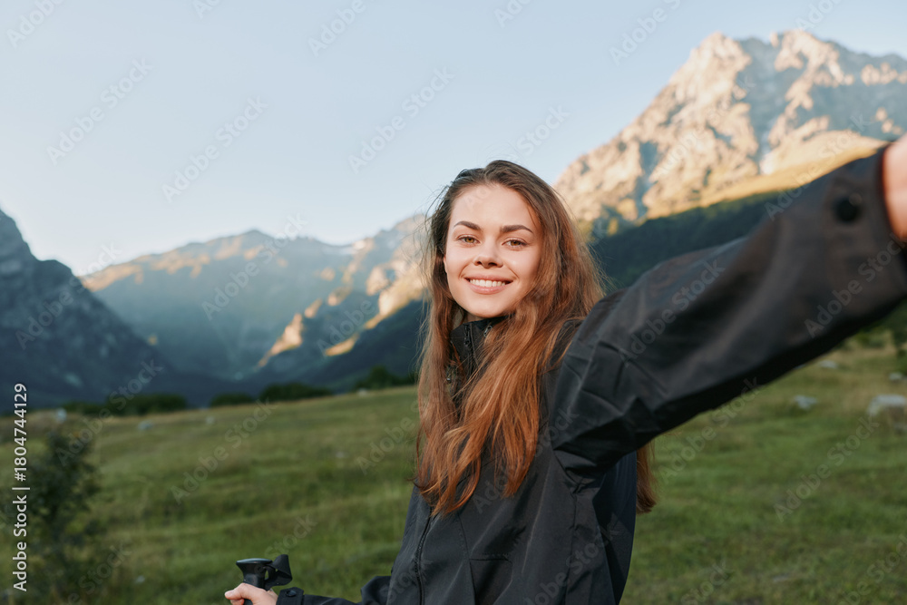 Fototapeta premium A cheerful young woman stands in a scenic mountain meadow, smiling as she takes a selfie with a compact camera, capturing a bright and energetic outdoor moment.