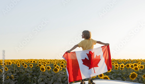 A child stands against the backdrop of a blooming sunflower field, holding a large Canadian flag behind his back. The country's national symbol. Happy Canada Day. Pride, patriotism