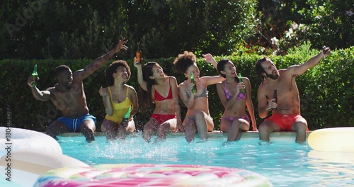 Clinking bottles, six friends sitting on backyard pool edge in swimwear, splashing near donut float