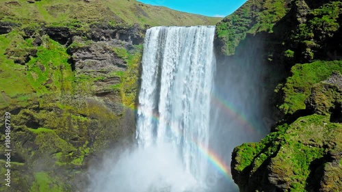 Aerial view of Skogafoss waterfall with rainbow forming in the mist. Drone footage capturing powerful white water plunging into the canyon below steep mossy cliffs.
