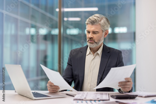 Mature businessman studying important paperwork, comparing financial reports and data analysis, making focused decisions while working in a modern corporate office