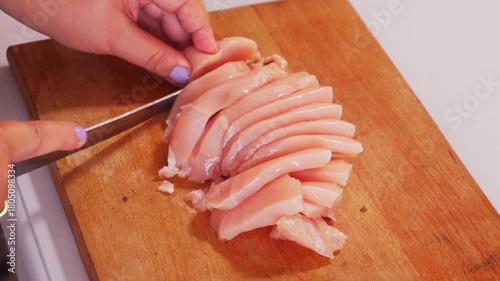 A woman's hands cut a chicken fillet into pieces on a cutting board on a table. Medium shot.