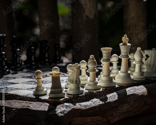 Close-up view of white chess pieces on a rustic outdoor chessboard. Strong directional light creates dramatic shadows and an atmospheric mood. Ideal for themes of strategy, focus.