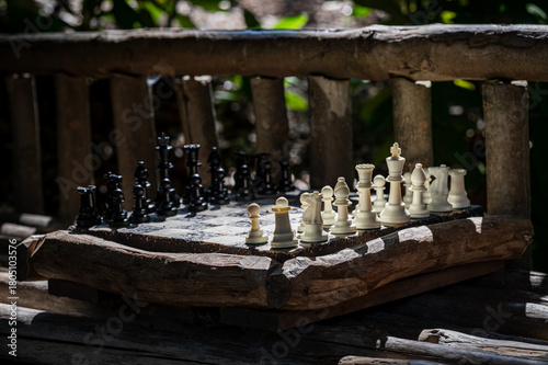 A stylized outdoor chessboard made of rustic wood with black and white chess pieces set for play. Sunlight creates dramatic contrast and a natural atmosphere in a forest-like setting. 