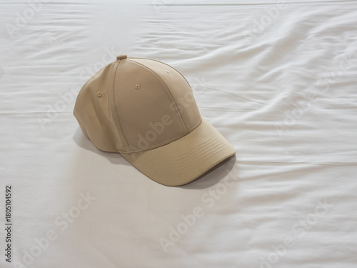A simple, light beige baseball cap rests casually on a gently rumpled white fabric background, offering a clean, minimalist aesthetic with soft, inviting lighting.