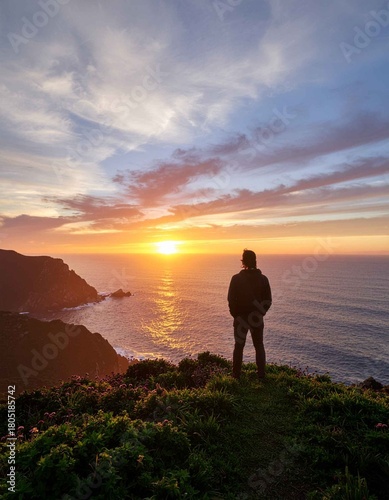 Lone traveler standing on a green coastal cliff, watching the vibrant and peaceful sunset over the ocean