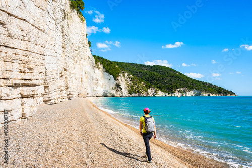 Female hiker walking along the pebble beach beneath the towering limestone cliffs of Vignanotica, Apulia, Italy