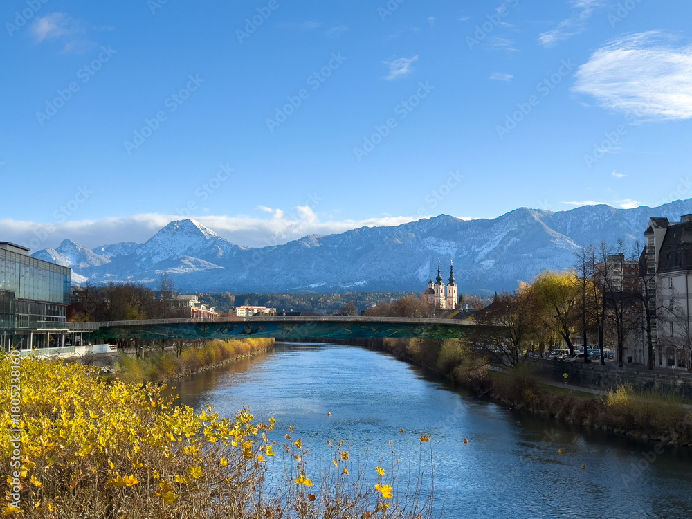 Fototapeta premium Villach, Austria - November 18, 2025: Scenic view of the river with autumn foliage, mountains in the background, and a historic church, capturing the beauty of nature and architecture