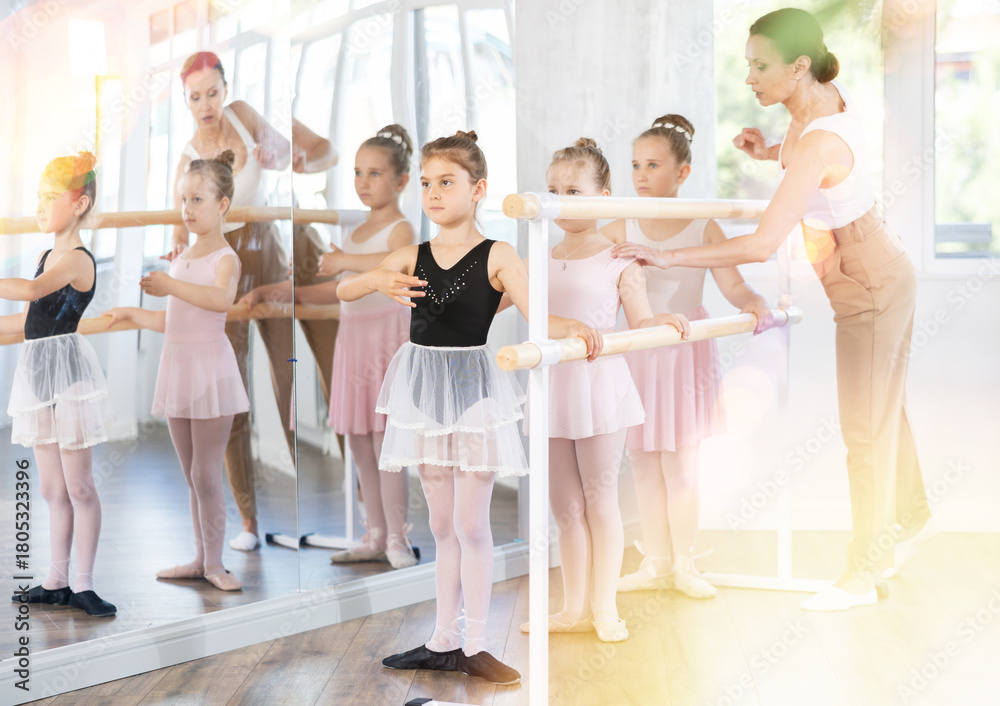 Naklejka premium Adult female teacher corrects group of girls dancers standing in first position at ballet barre