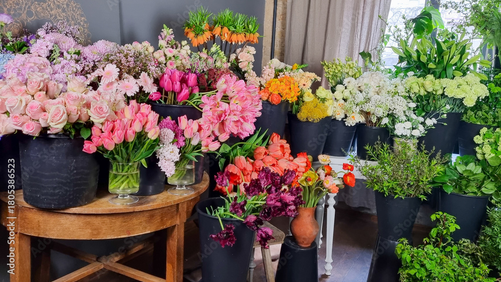 Fototapeta premium Colorful display of fresh flowers arranged in black buckets inside a florist shop. A variety of vibrant blooms pink tulips, roses, white flowers, greenery, and mixed seasonal plants in flower shop