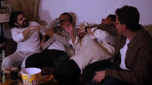 four young Hispanic men on a couch. Three friends are laughing and gesturing at the fourth, who appears embarrassed or annoyed. Highlights social teasing and shared home entertainment.