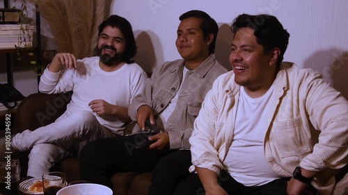 three young Hispanic men on a couch, laughing and talking while enjoying a show or movie. Highlights shared humor, social interaction, and home entertainment
