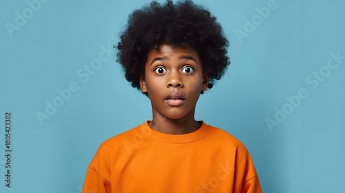 Vibrant studio portrait of a young child with voluminous afro hair wearing a bright orange shirt against a smooth blue backdrop, captured with crisp detail and expressive, engaging presence