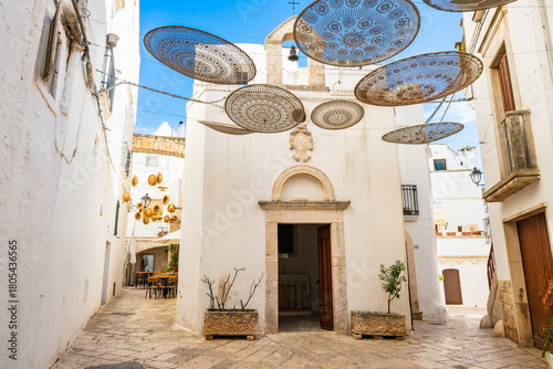 Fototapeta Naklejka Na Ścianę i Meble -  Charming narrow street with lace-patterned parasols hanging above a small white chapel in Locorotondo town, Apulia, Italy