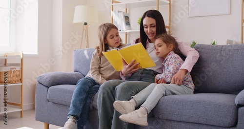 Happy family at home, mother or babysitter holding read book, embrace cute little children daughter, son, parent mum tells small kids funny fairy tale story, sit on sofa having fun together at home