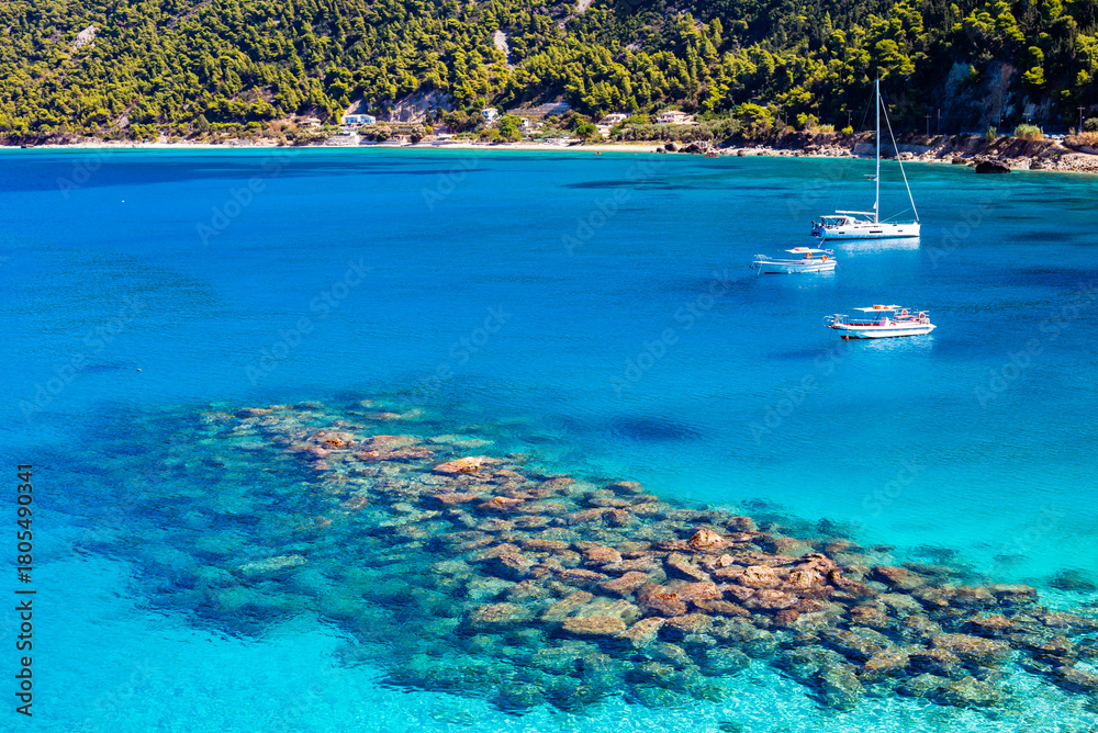 Fototapeta premium Rocks and stones in crystal clear turquoise sea water of Agios Nikitas bay with sailing boats, Lefkada island, Greece