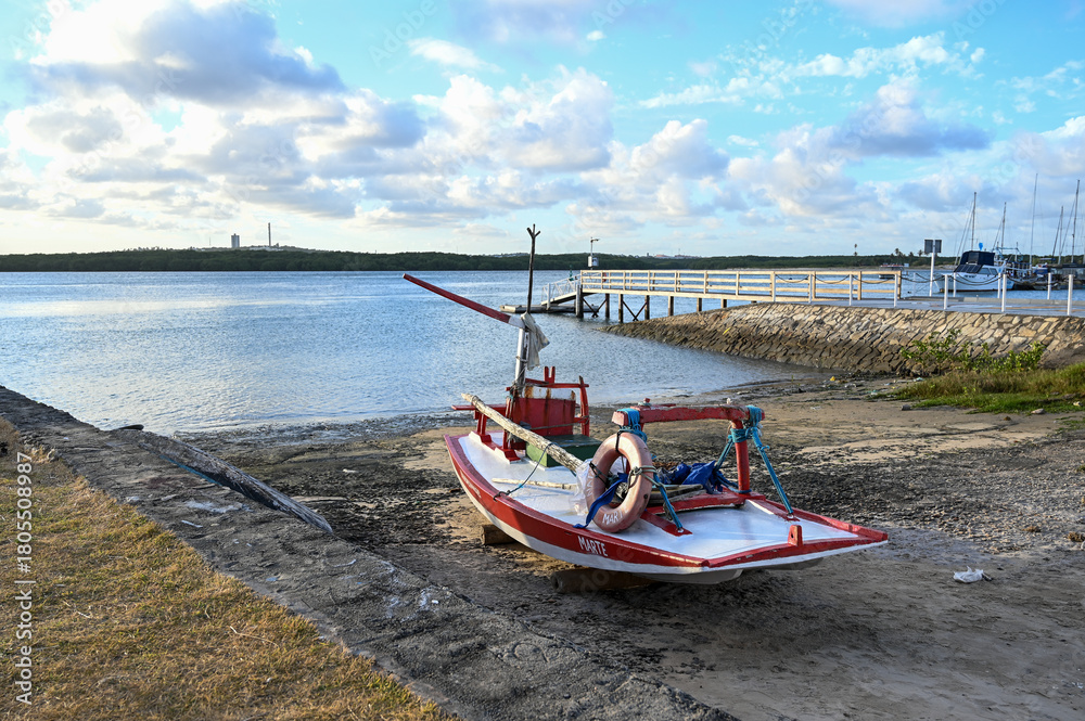 Obraz premium small fishing boat on dry land, potengi river natal brazil, local maritime scene traditional red and white boat, boat ramp and pier, cloudy sky landscape vessel on shore at low tide, coastal area