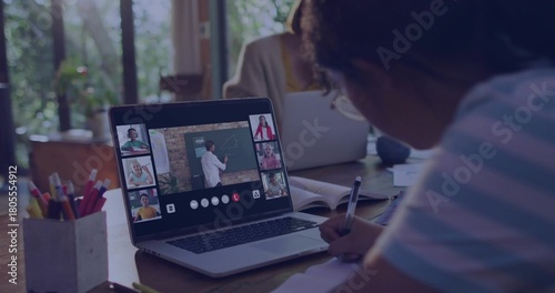 Writing child in striped shirt sitting at table holding pen over notebook watching laptop call
