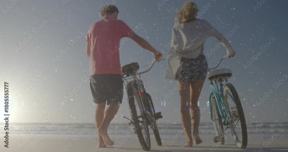 Naklejka premium Walking couple wearing cover-up and pink tee pushing two bicycles on wet sand, low sun reflecting