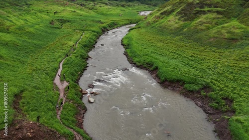 Clear mountain water flows over rocks in a vibrant green valley, surrounded by majestic peaks at dawn.
